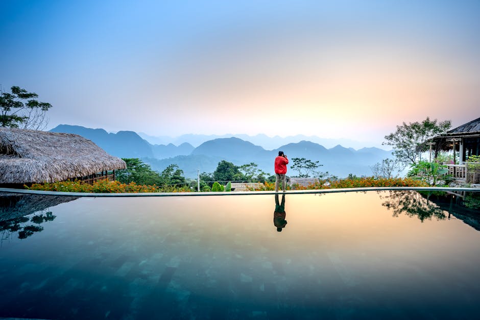 Anonymous distant tourist standing on edge of swimming pool on terrace near wooden houses against mountainous terrain in evening time