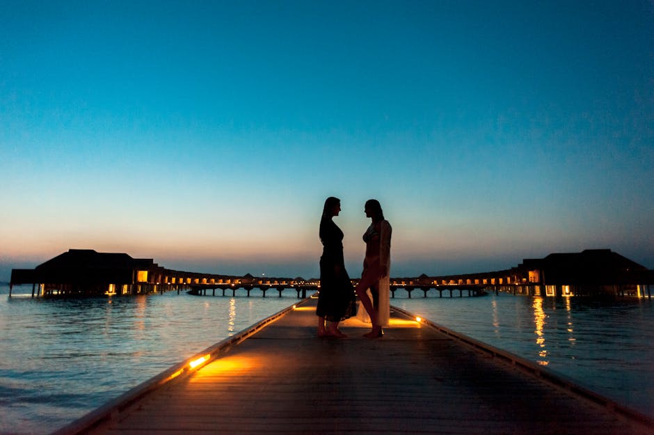 Two silhouetted women sharing a romantic moment on a Maldives boardwalk at dusk